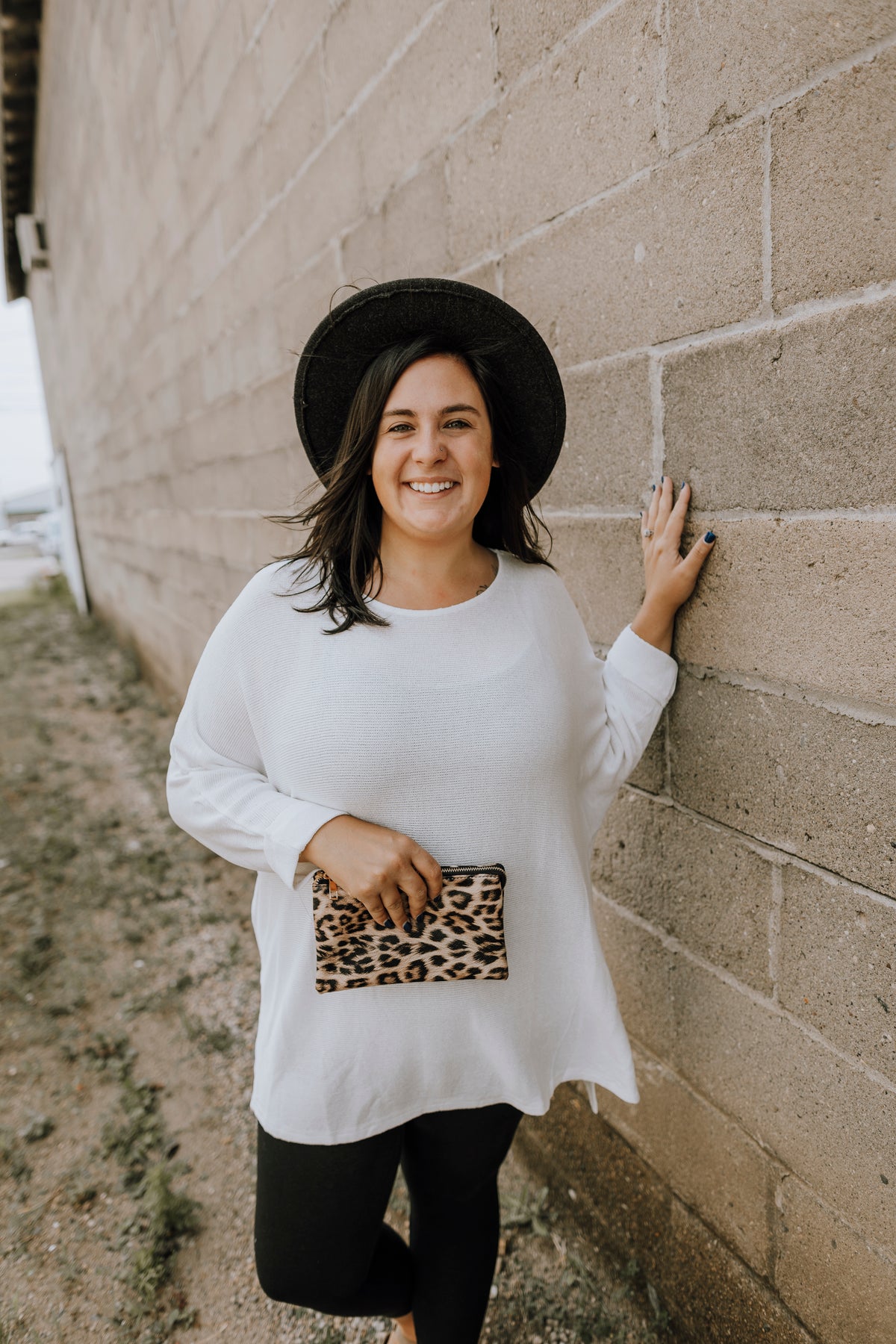 Anna has dark brown hair and is wearing a plus size slouchy ivory high low 3/4 sleeve sweater in size 1X with a charcoal wide brim hat, leopard clutch and black leggings.