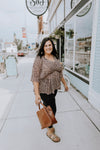 Anna has dark brown hair and is wearing a plus size dusty peach leopard drop shoulder empire waist top in size 1X with black jeans and a cognac brown handbag.