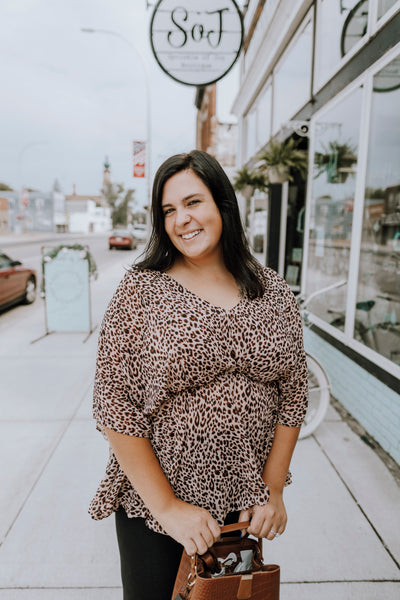 Anna has dark brown hair and is wearing a plus size dusty peach leopard drop shoulder empire waist top in size 1X with black jeans.